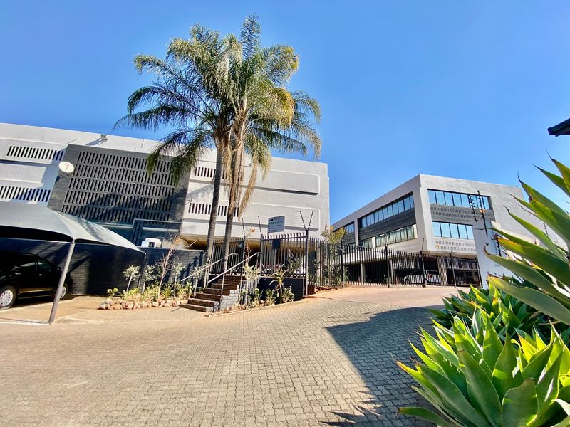 Front exterior view of a modern commercial office complex featuring paved driveway, secure gated access, electric fencing, and covered parking bays.