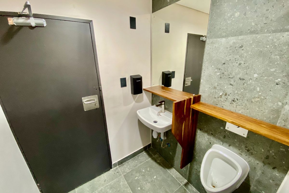 A modern commercial restroom featuring a wall-mounted basin, urinal, wooden vanity shelf, and large mirror with contemporary grey tiled finishes.