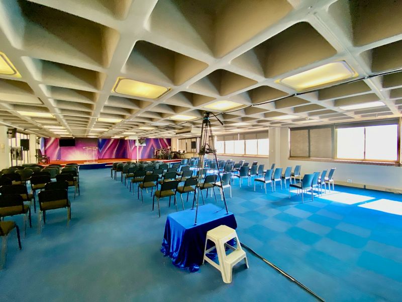 A large commercial assembly hall featuring a distinctive waffle slab ceiling, raised stage area, and ample natural light from large windows.