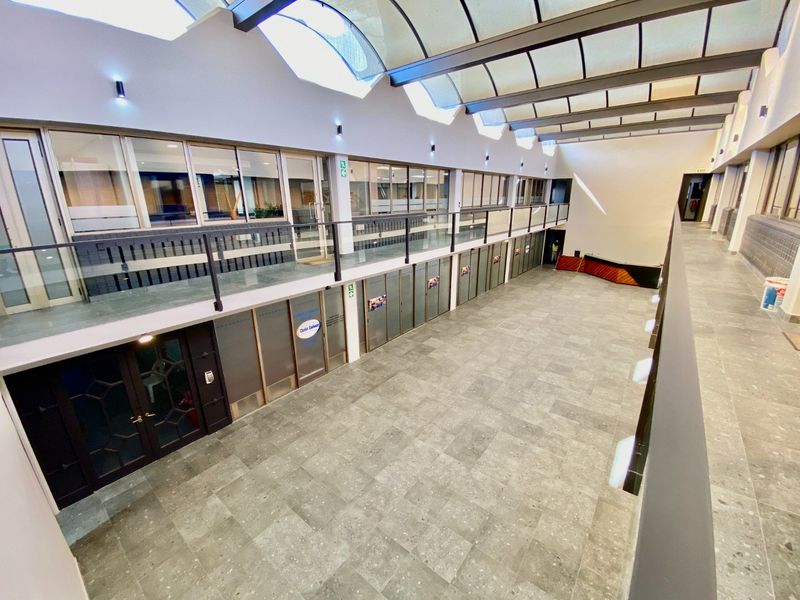 Spacious commercial atrium hallway featuring a high glass roof, mezzanine level with glass balustrades, and modern tiled flooring throughout.
