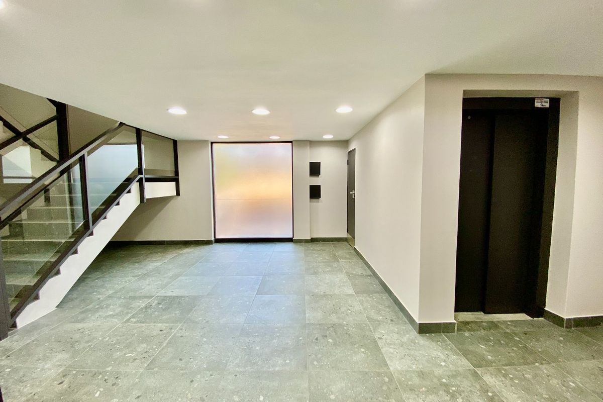 A modern, minimalist commercial building entrance lobby featuring tiled flooring, a sleek elevator, and a staircase with a glass balustrade.