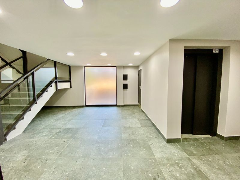 A modern, minimalist commercial building entrance lobby featuring tiled flooring, a sleek elevator, and a staircase with a glass balustrade.