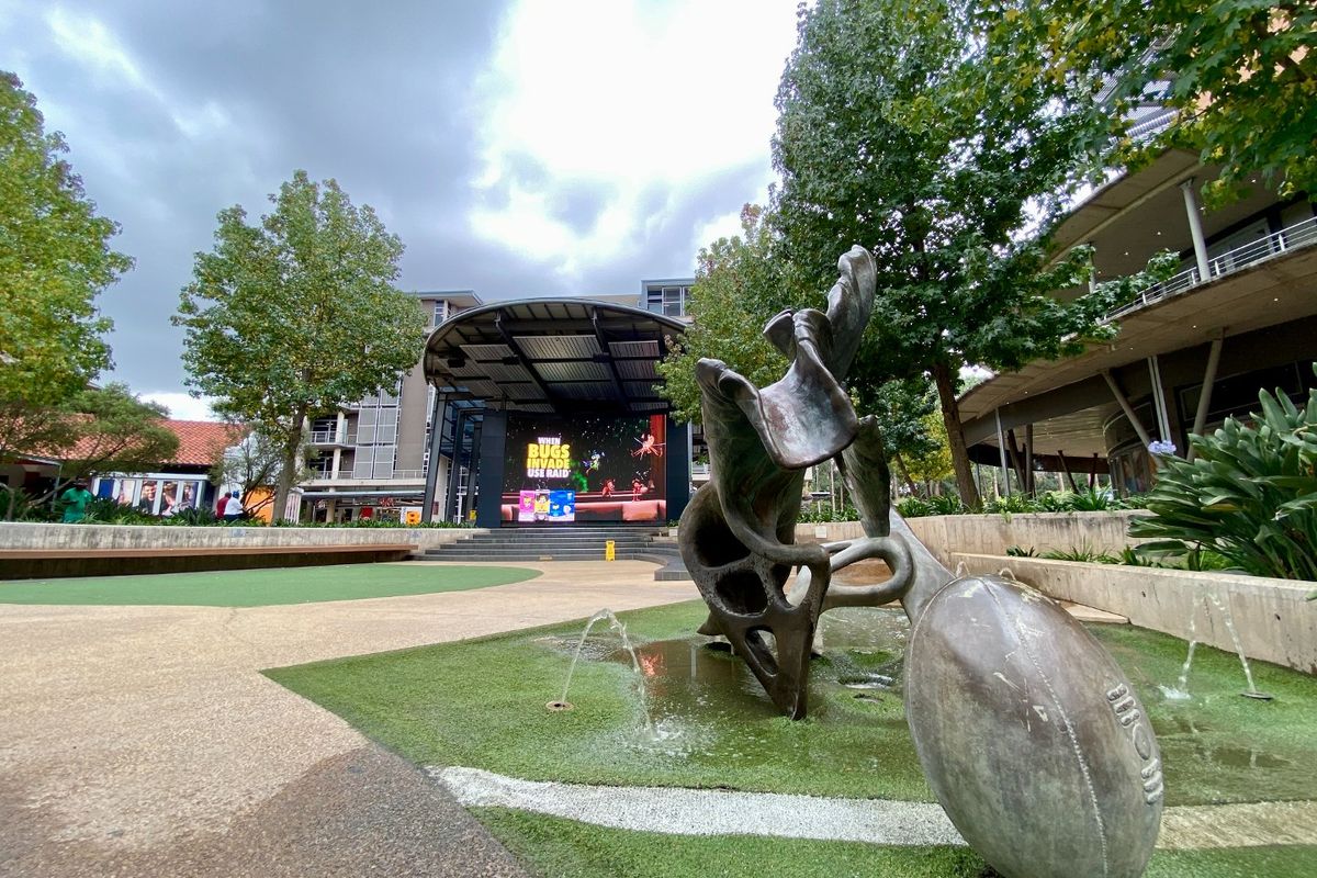 A well-maintained commercial outdoor plaza featuring a stage with a large digital screen, a sculptural water feature, and landscaped paved areas.