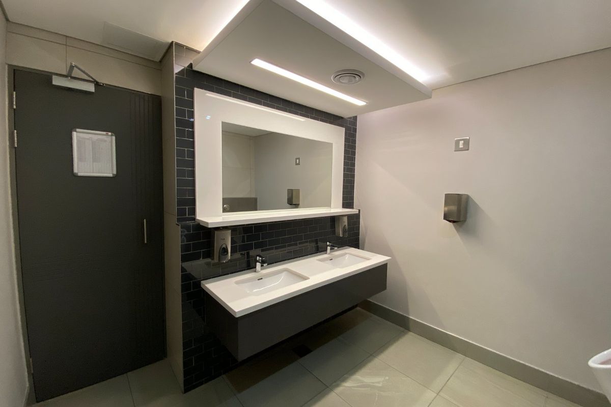 Modern commercial bathroom featuring a sleek double vanity with integrated mirror, subway tile backsplash, and recessed ceiling lighting.