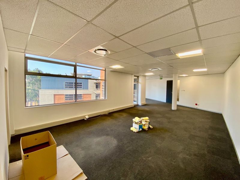 An open-plan office space featuring a suspended ceiling, recessed lighting, and large windows providing natural light and views of the surrounding commercial complex.