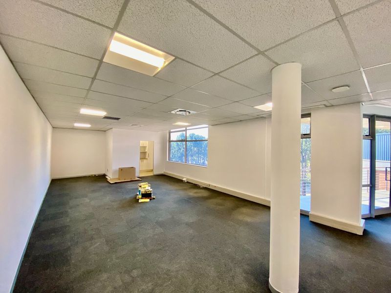 An open-plan commercial office space featuring a suspended ceiling, recessed lighting, carpet tiles, and a structural column with large windows providing natural light.