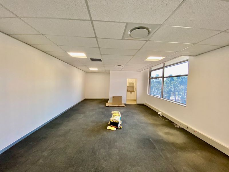 An empty, open-plan commercial office space featuring a suspended ceiling, recessed lighting, large windows, and an adjoining kitchenette area.