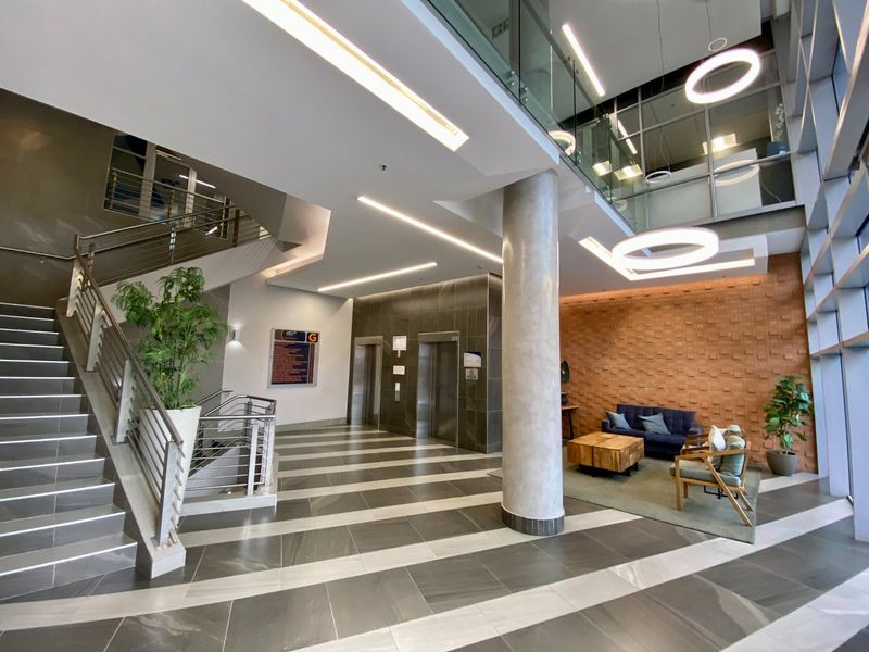 Modern commercial building lobby featuring a double-volume entrance, elevator access, a feature staircase with glass balustrades, and contemporary tiled flooring.