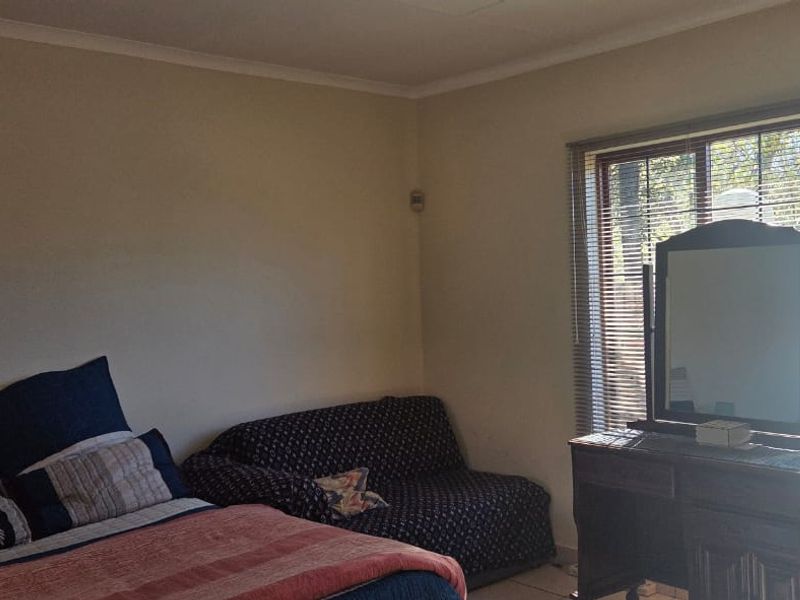 A bedroom featuring neutral walls, tiled flooring, and large windows fitted with venetian blinds and a security alarm sensor.