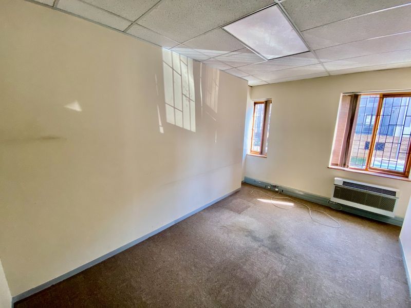 An empty commercial office space featuring a suspended ceiling, wall-mounted HVAC unit, and carpeted flooring with visible wear.
