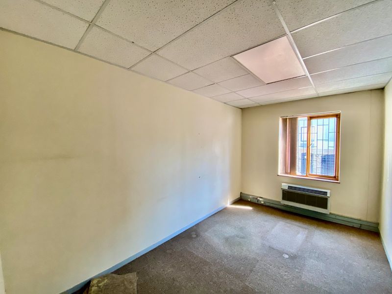 An empty commercial office space featuring a suspended ceiling, recessed lighting, a window with security bars, and a wall-mounted HVAC unit.