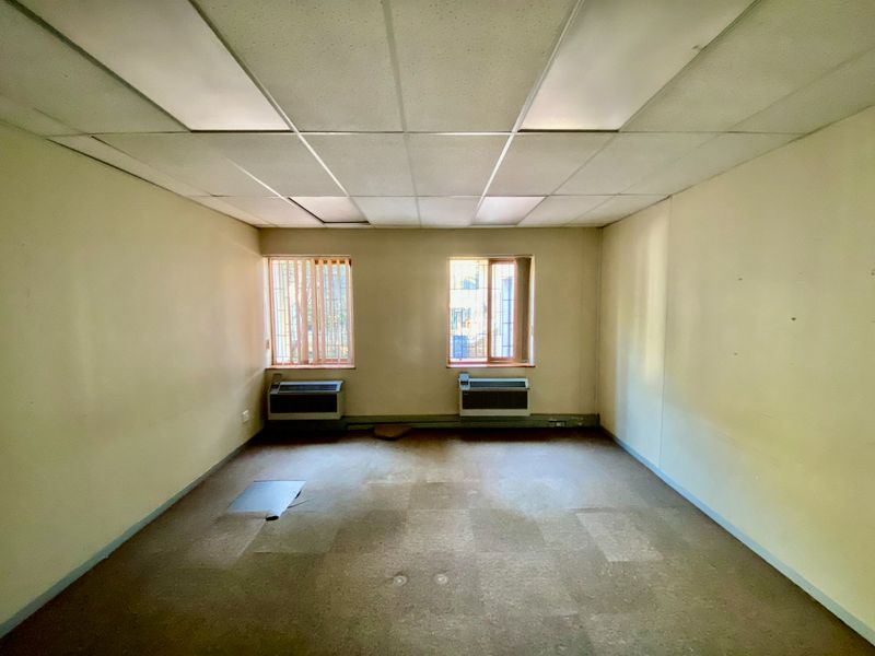 An empty commercial office space featuring a suspended ceiling, recessed lighting, and two PTAC air conditioning units with security bars on the windows.