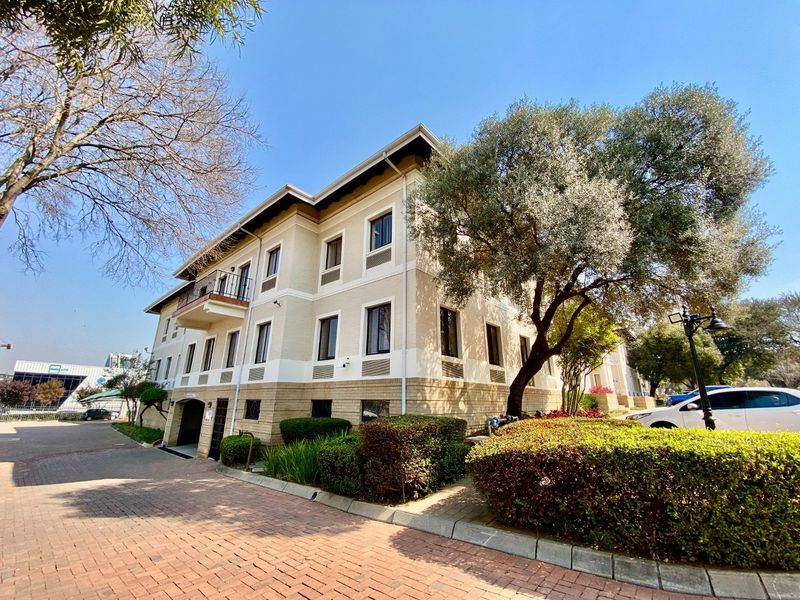 A well-maintained multi-story commercial building featuring a brick-paved driveway, landscaped garden areas, and a balcony on the upper level.