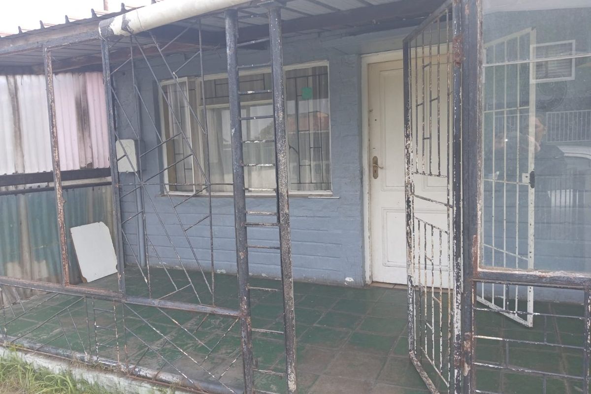 A covered patio area featuring a metal security gate, tiled flooring, and a corrugated roof structure, showing signs of wear and weathering.