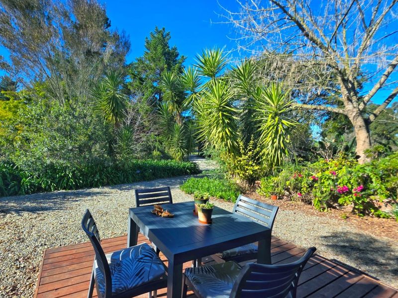 A timber deck patio area set within a lush, landscaped garden featuring mature trees and gravel pathways.