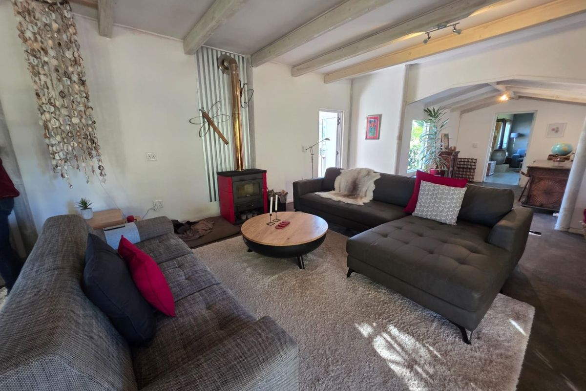 A cozy living room featuring exposed ceiling beams, a wood-burning stove with corrugated metal backing, and direct access to adjacent living spaces.