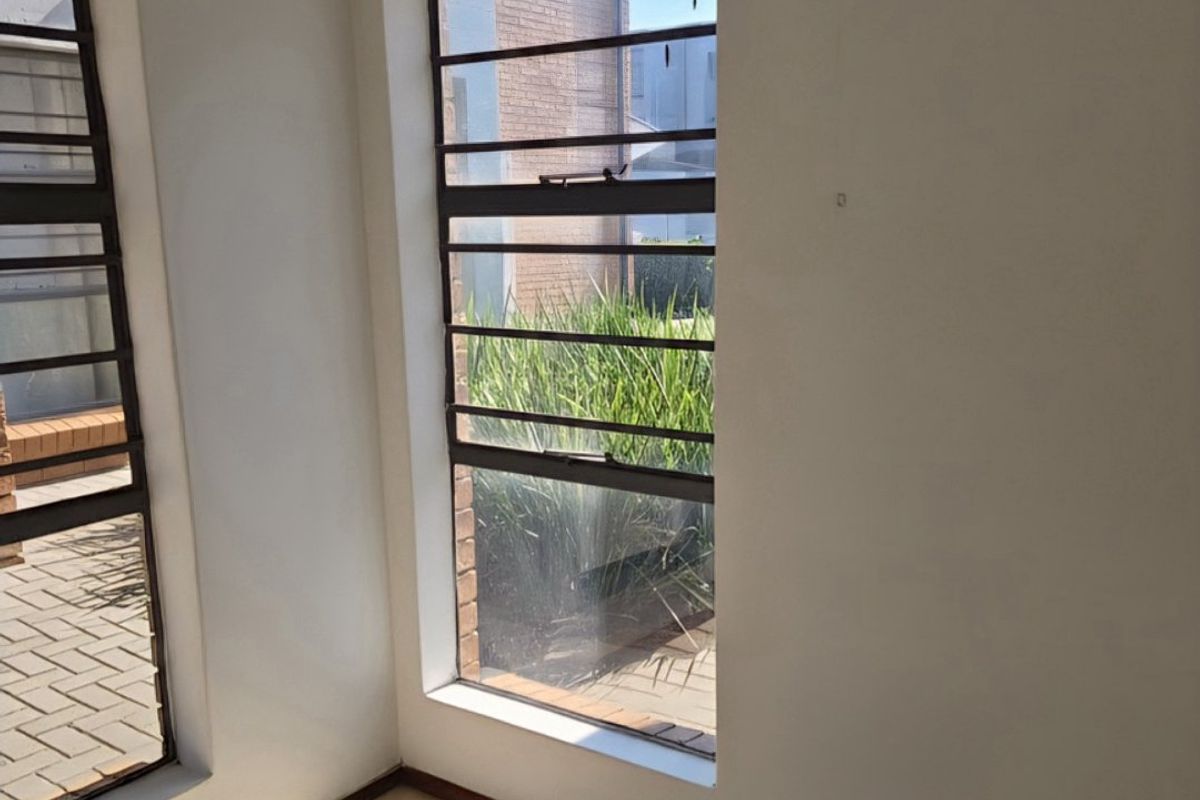 An empty living area featuring neutral tiled flooring, large windows with security bars, and curtain tracks, offering a bright and blank canvas.