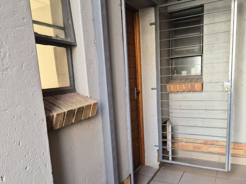 A small exterior balcony or service area featuring tiled flooring, a security gate, and a downpipe, showing signs of wear on the exterior walls.