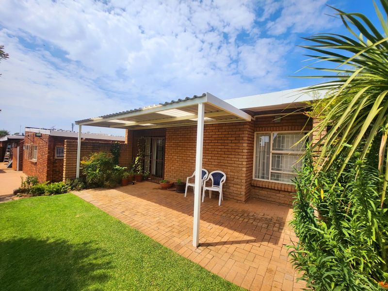A covered brick-paved patio area featuring a corrugated roof structure, security gate, and burglar bars, overlooking a small garden space.