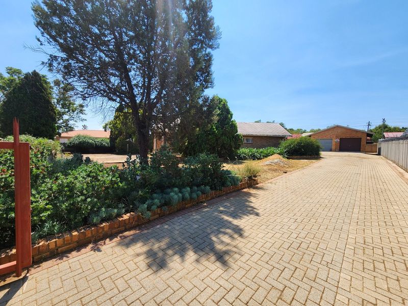 Front view of the property featuring a long paved driveway, established garden beds with brick retaining walls, and a detached garage structure.