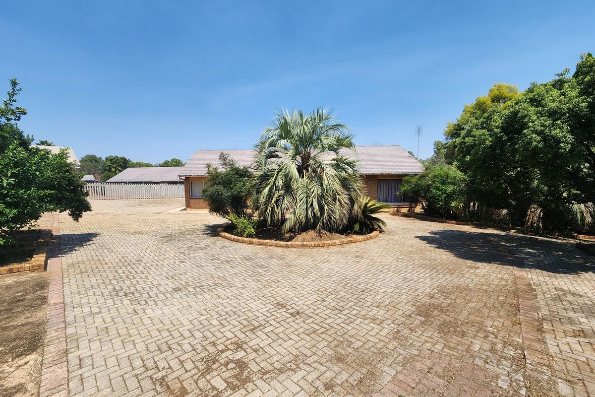 A spacious front exterior featuring a large paved driveway, a central landscaped garden bed, and a brick house facade with a tiled roof.
