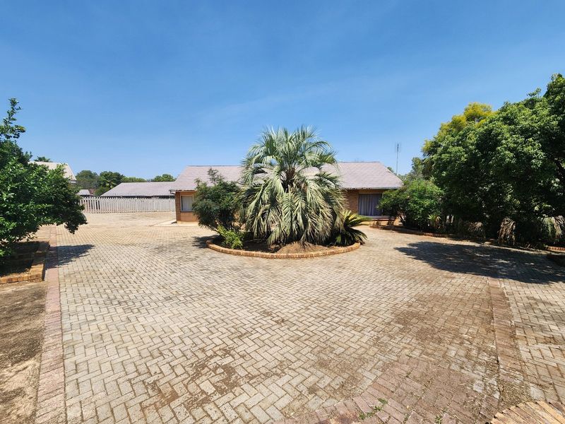 A spacious front exterior featuring a large paved driveway, a central landscaped garden bed, and a brick house facade with a tiled roof.