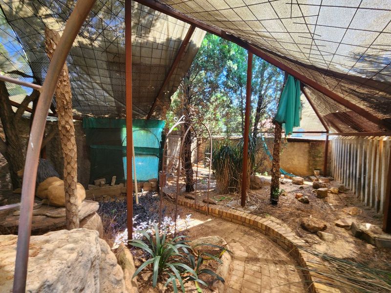An outdoor patio area featuring a metal-framed shade cloth canopy, brick-edged pathways, and established garden landscaping.