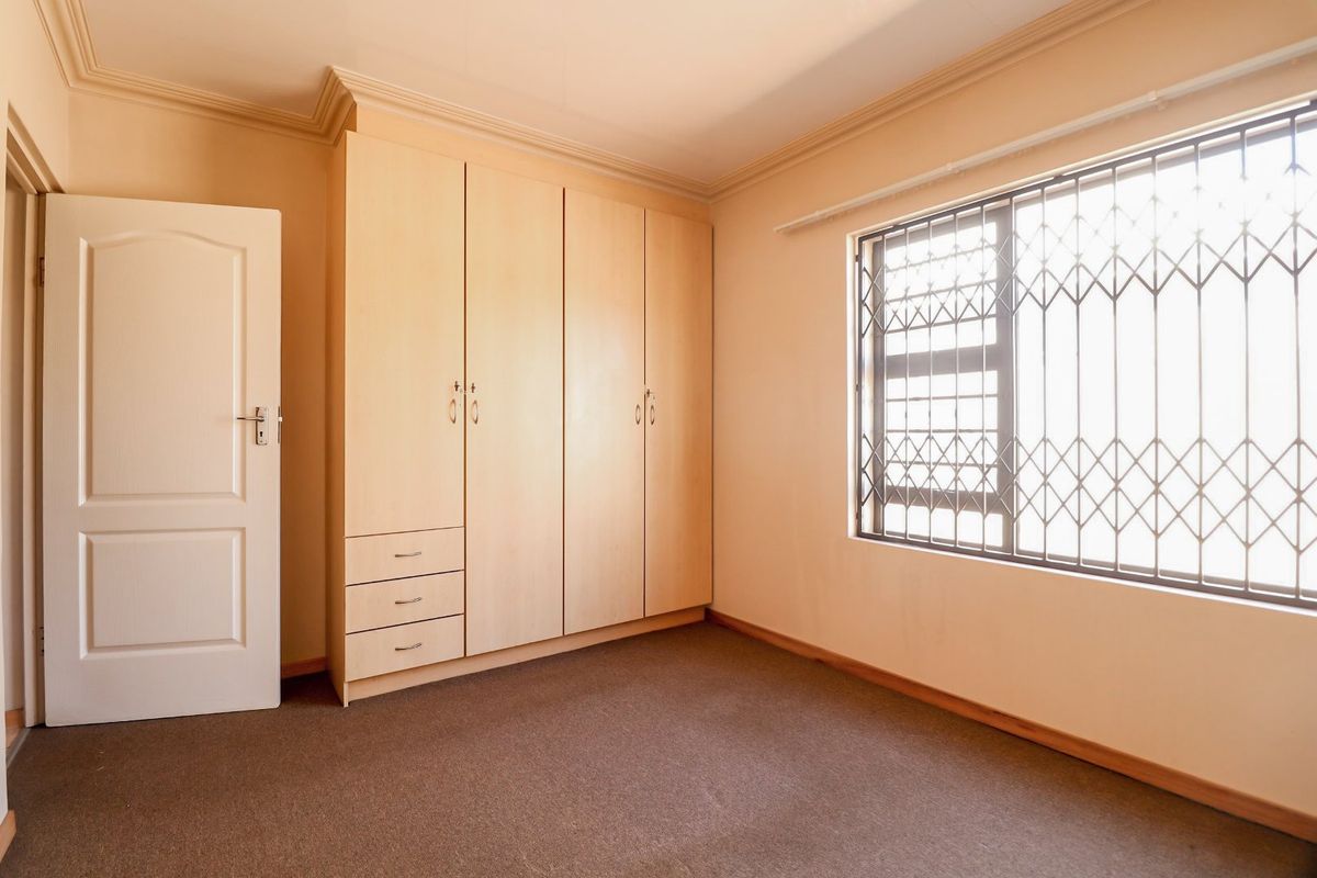 A clean, empty bedroom featuring built-in wooden cupboards, carpeted flooring, and a window fitted with a security gate.