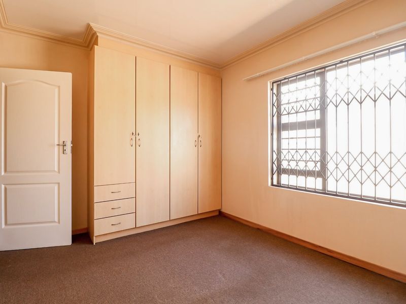 A clean, empty bedroom featuring built-in wooden cupboards, carpeted flooring, and a window fitted with a security gate.