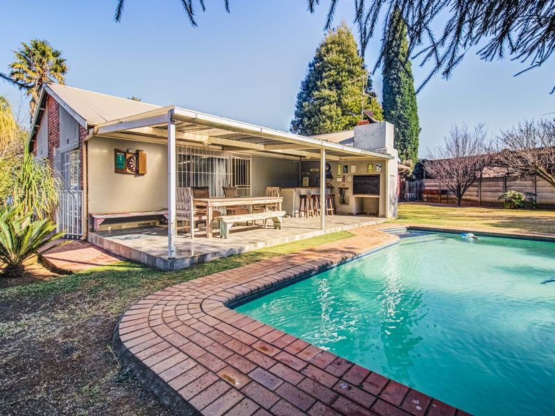 Outdoor entertainment area featuring a covered patio with a built-in braai, overlooking a swimming pool with brick coping in a landscaped garden setting.