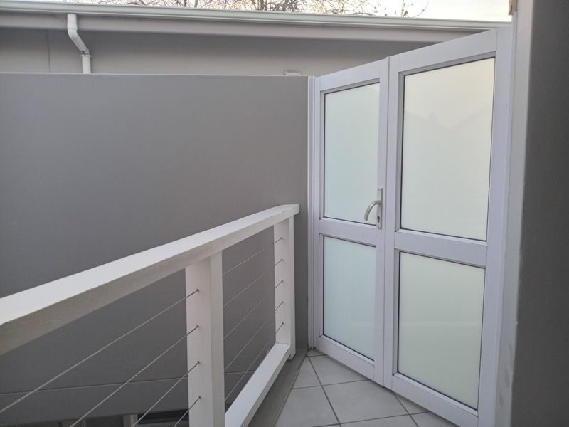 A clean, modern balcony area featuring tiled flooring, a white metal safety railing, and a frosted glass door providing access to the interior.