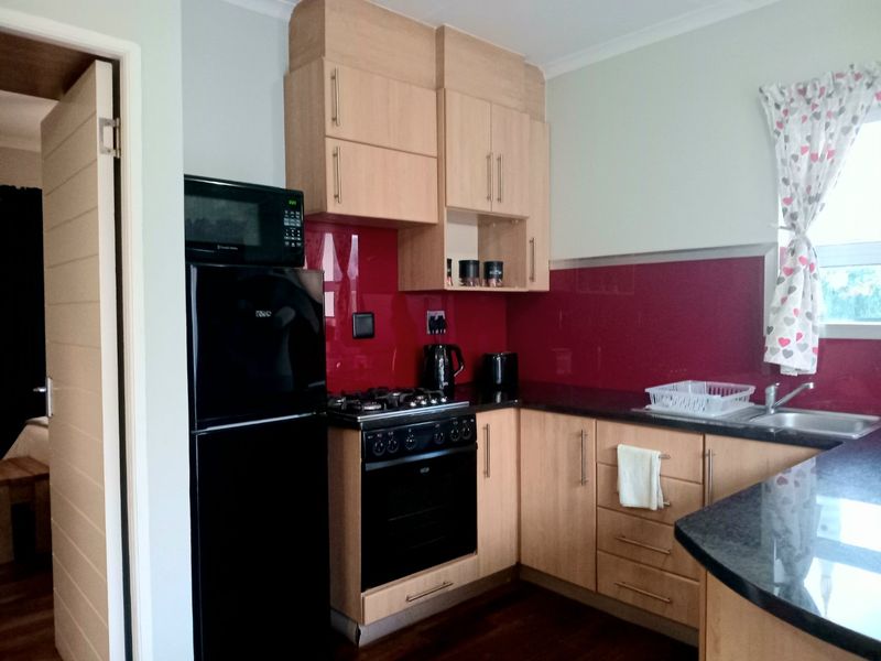 A functional kitchen featuring light wood cabinetry, dark granite countertops, a bold red glass splashback, and integrated appliances including a gas hob and oven.