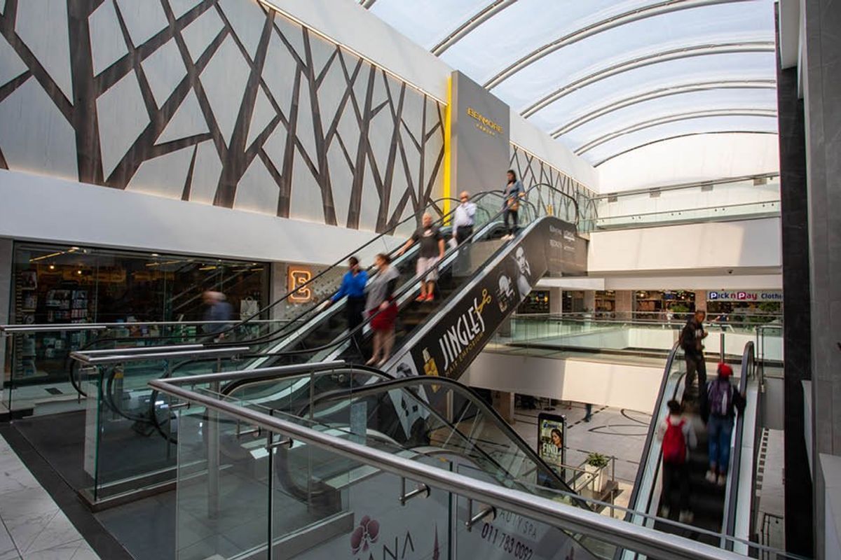 Modern commercial shopping center atrium featuring multiple escalators, glass balustrades, and a large curved skylight providing natural illumination.