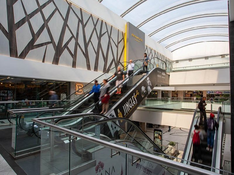 Modern commercial shopping center atrium featuring multiple escalators, glass balustrades, and a large curved skylight providing natural illumination.