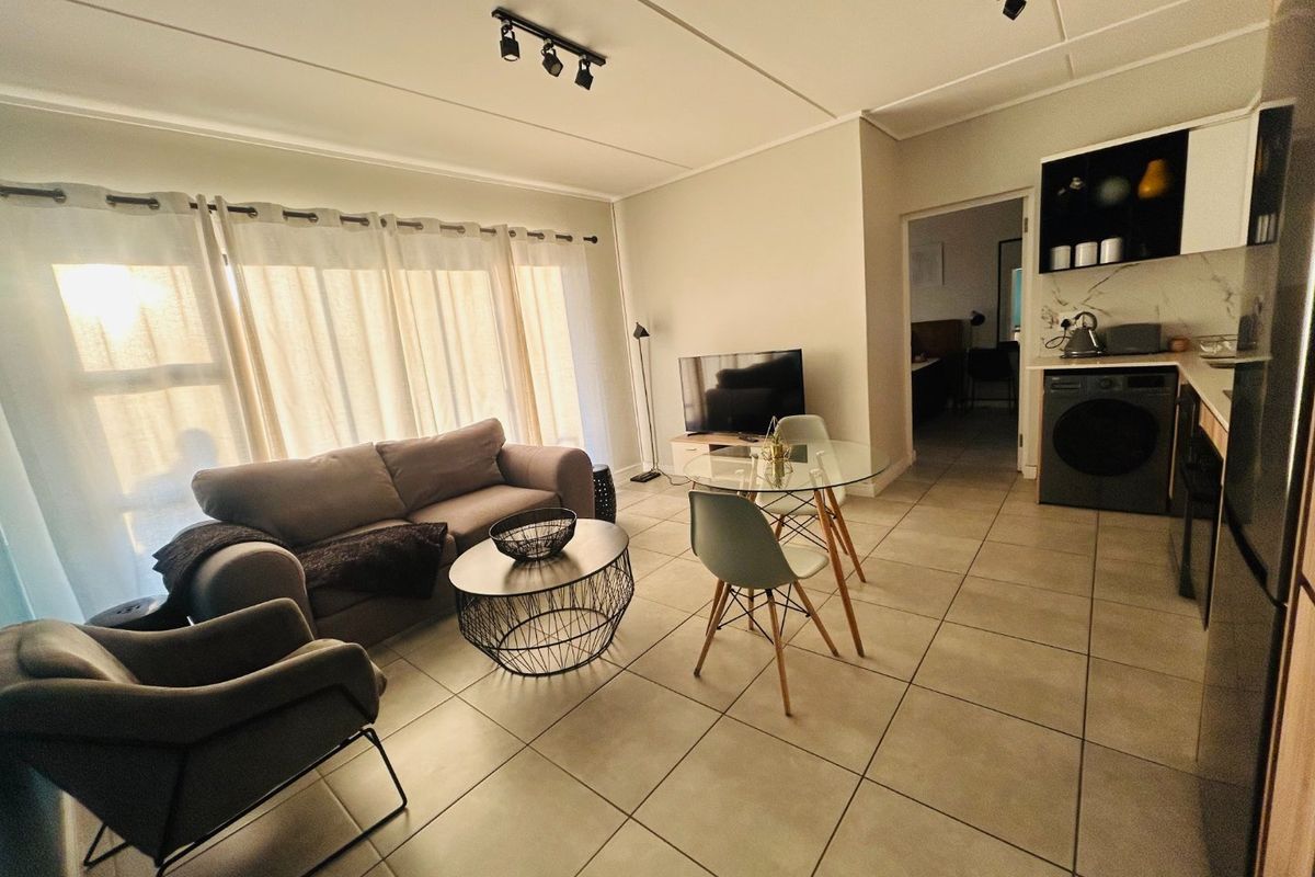 An open-plan living and kitchen area featuring neutral tiled flooring, modern track lighting, and a compact, functional kitchen layout with built-in cabinetry.