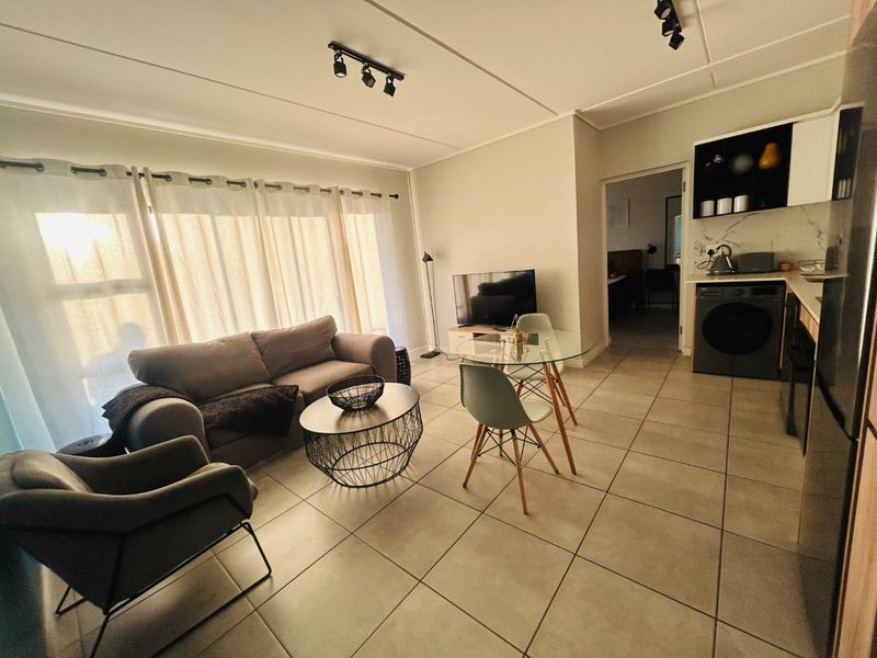 An open-plan living and kitchen area featuring neutral tiled flooring, modern track lighting, and a compact, functional kitchen layout with built-in cabinetry.