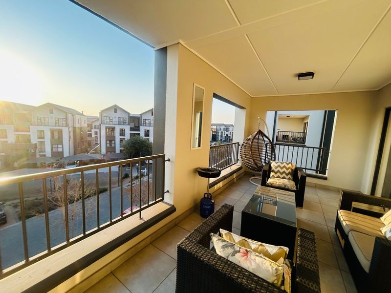 A spacious covered balcony featuring tiled flooring and a metal balustrade, offering views of the surrounding apartment complex.