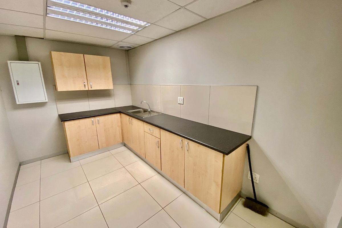A clean, functional commercial kitchenette featuring light wood-finish cabinetry, dark laminate countertops, a stainless steel sink, and neutral tiled flooring.