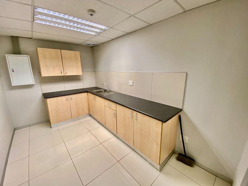 A clean, functional commercial kitchenette featuring light wood-finish cabinetry, dark laminate countertops, a stainless steel sink, and neutral tiled flooring.