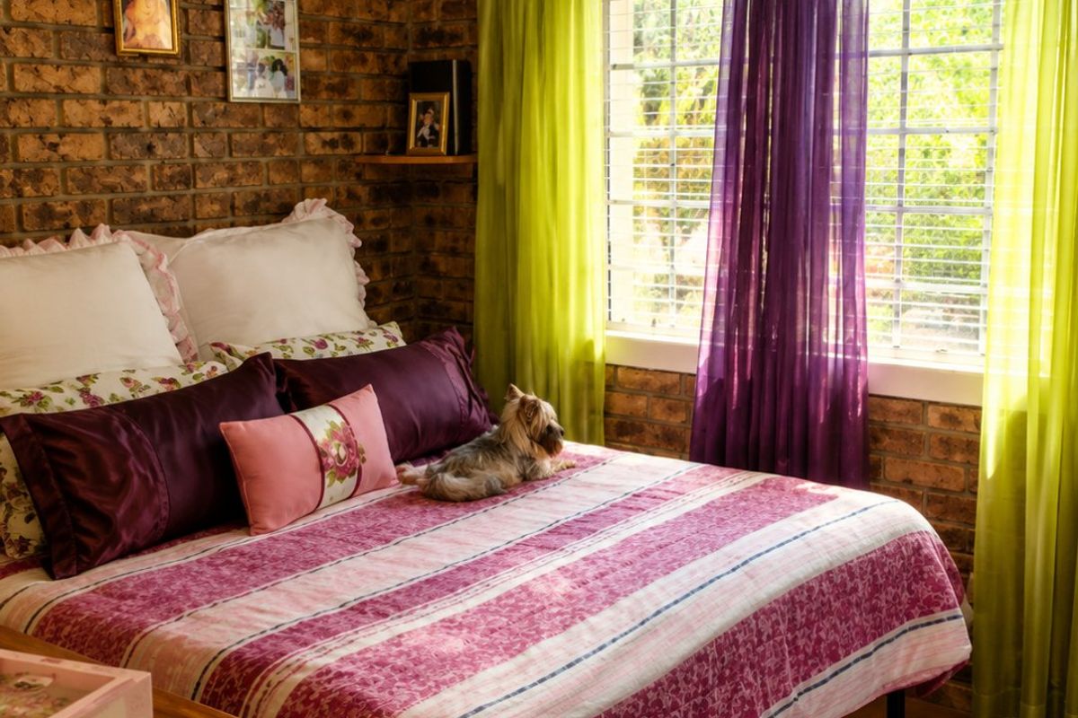 A bedroom featuring rustic exposed brick walls, tiled flooring, and a window fitted with security bars, offering a warm and textured aesthetic.