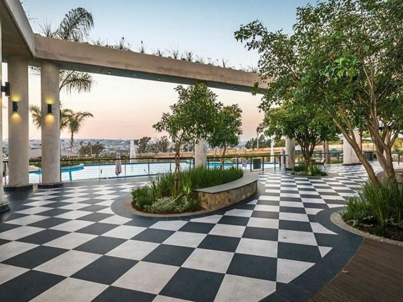 A well-maintained outdoor pool area featuring a striking checkered stone patio, concrete pergola structure, and glass balustrades overlooking the landscape.