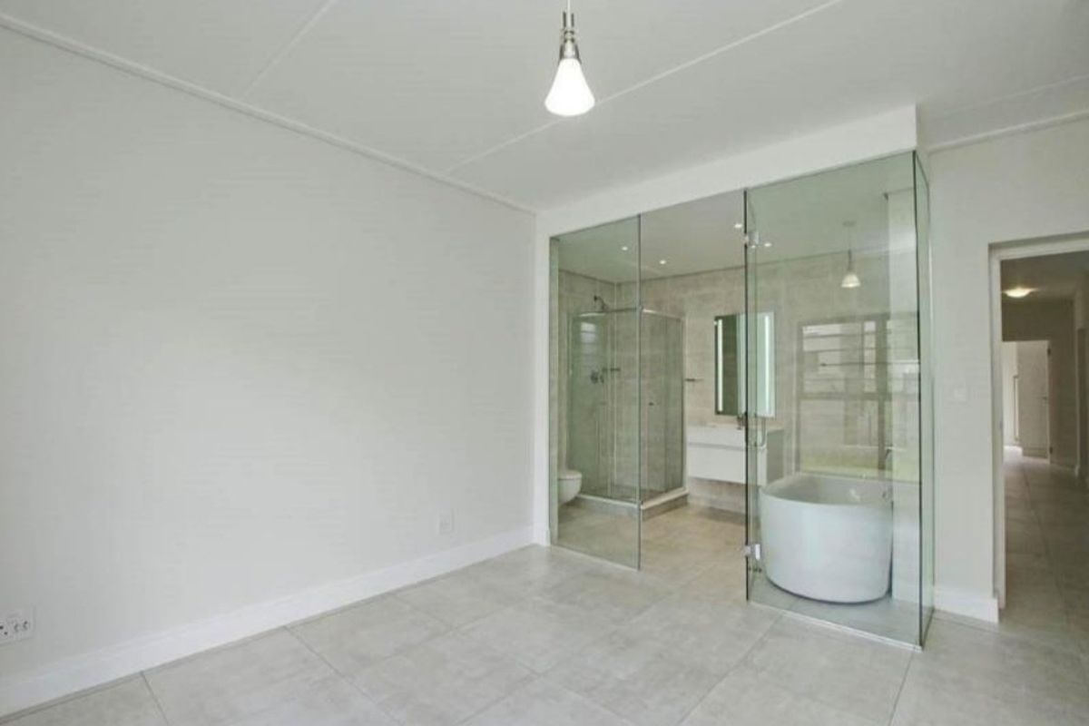 A modern, minimalist bedroom featuring an open-concept glass-enclosed ensuite bathroom with a freestanding tub, walk-in shower, and sleek wall-mounted vanity.