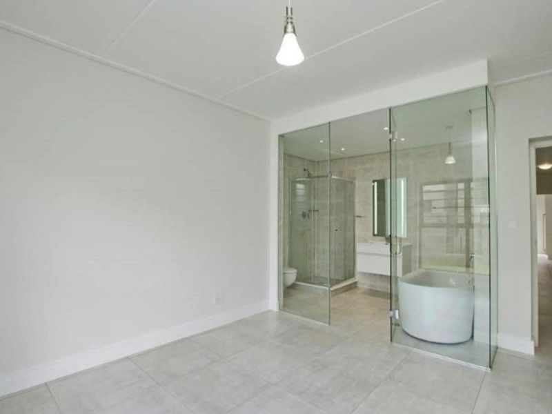 A modern, minimalist bedroom featuring an open-concept glass-enclosed ensuite bathroom with a freestanding tub, walk-in shower, and sleek wall-mounted vanity.