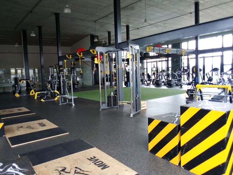 A spacious, industrial-style gym featuring exposed concrete ceilings, steel structural beams, and durable rubberized flooring with large windows providing ample natural light.