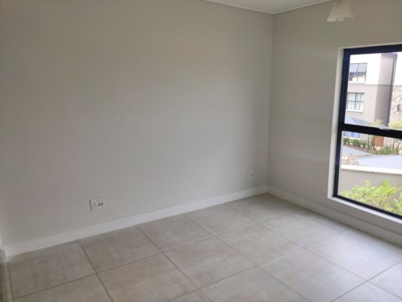 A bright, empty bedroom featuring neutral tiled flooring, clean walls, and a large window with a black aluminium frame.