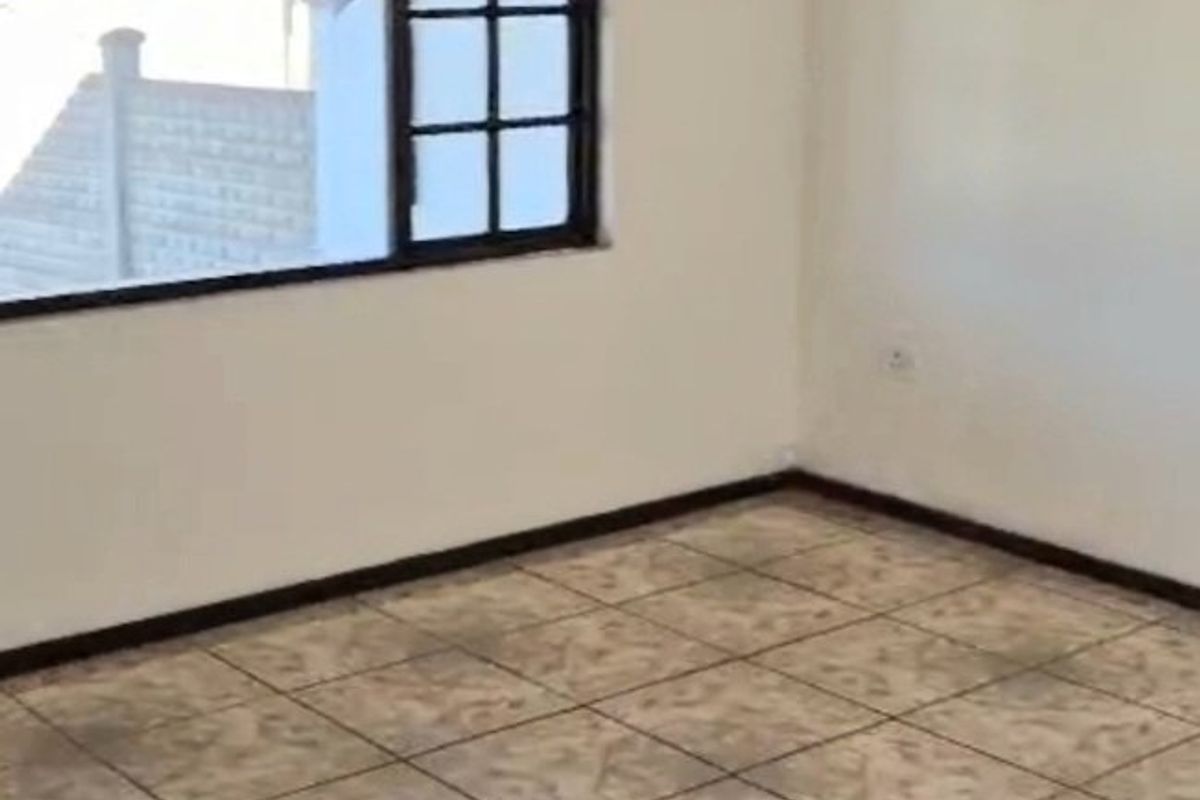 An empty living room featuring neutral ceramic floor tiles, white walls, and a large grid-pattern window providing natural light.