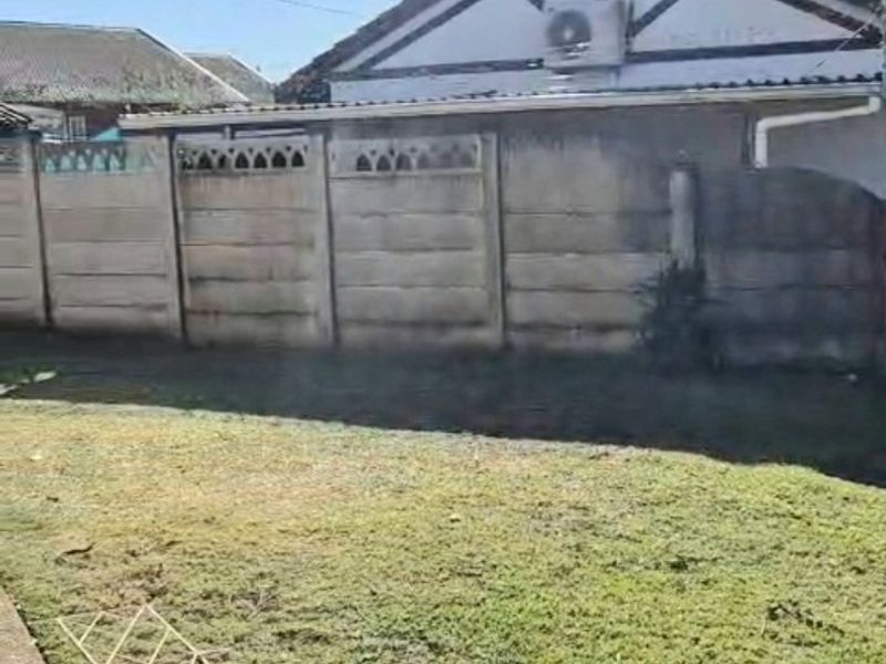 Rear exterior view of the property featuring a lawn, concrete pathway, and a precast concrete boundary wall with an external air conditioning unit.