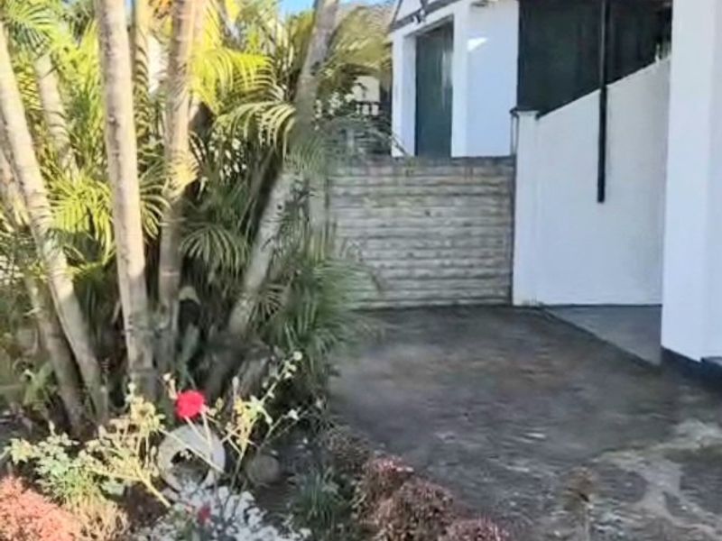 Front exterior view of the apartment featuring a paved driveway, established garden with palm trees, and a satellite dish mounted on the roof.