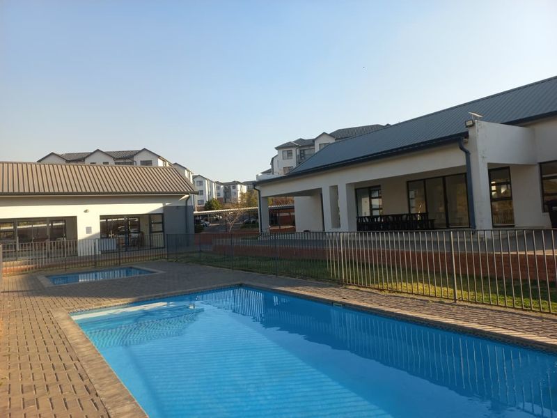 A well-maintained outdoor pool area featuring a paved deck, metal perimeter fencing, and an adjacent covered patio area within a residential complex.