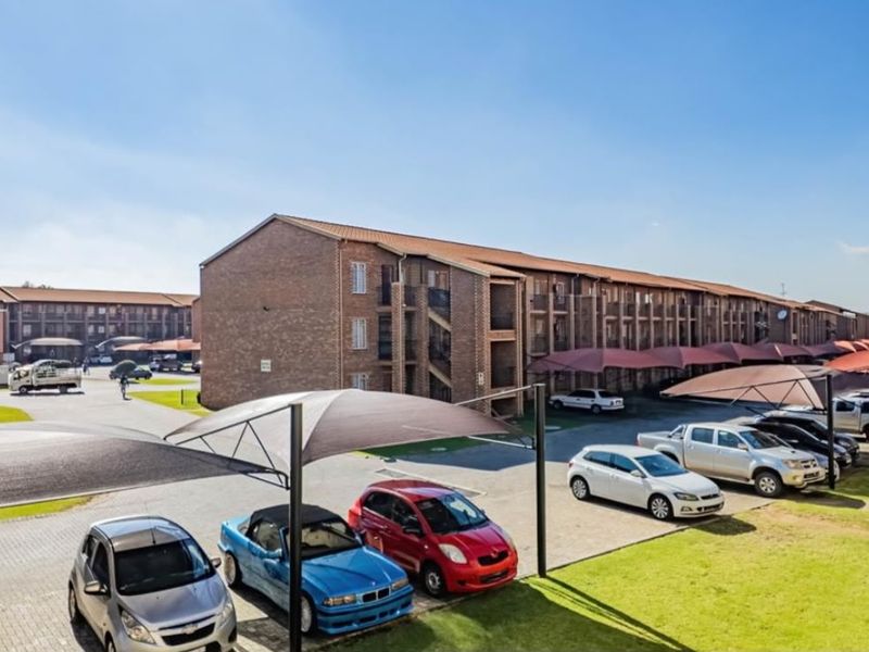 Exterior view of a multi-story brick apartment complex featuring paved parking areas with shade cloth carports and surrounding landscaped grounds.